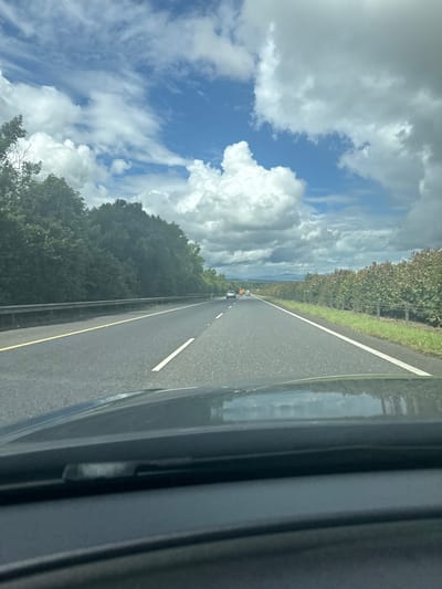 This media file depicts a view from inside a vehicle traveling on a rural road. The road is paved and has a white line marking the center and a yellow line marking the shoulder on the right side. A dark-colored vehicle is visible in the distance on the road, suggesting a traveling activity.
The scene is set outdoors, with lush green trees lining the left side of the road. Beyond the trees, a vast expanse of blue sky with dramatic, fluffy white clouds dominates the view. The clouds are a prominent feature, suggesting potentially dynamic weather conditions or simply a visually striking sky. A hint of a distant landscape, possibly hills or mountains, can be seen on the horizon behind the trees.
The right side of the image shows the interior of the vehicle, specifically a dark, textured surface that appears to be part of the car's interior trim, possibly the dashboard or door panel. Reflections of the road and sky are visible on this surface, adding to the impression of movement and the surrounding environment.
The lighting suggests it is daytime, possibly late morning or afternoon, given the brightness of the sky and the shadows cast. The overall mood conveyed is one of travel and observation of the natural landscape. There is no visible text in the image.