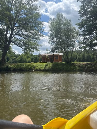 A view from a kayak, partially seen on the right, over a calm, murky river. The opposite bank is lush with trees and vegetation, leading to a small wooden house with a porch visible through the foliage. The sky above is filled with clouds, and trees frame the left side of the image. The overall scene evokes a sense of tranquility and immersion in nature.