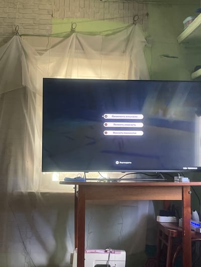 The image shows a television screen displaying a menu with several options. The menu is in Russian and features three selectable options: "Продолжить испытание" (Continue trial), "Понизить сложность" (Lower difficulty), and "Покинуть подземелье" (Leave dungeon). There is also a "Подтвердить" (Confirm) button. The screen is displaying a game interface, likely from an RPG or adventure game.
The television is placed in a room with a light green wall and a brick wall visible on the left side of the image. A white translucent fabric is draped in front of the brick wall, creating a soft, diffused light. The room appears to be somewhat cluttered, with some items visible on a wooden shelf unit to the right of the TV. One of these items is a white electronic device with blue lights. The overall impression is of an indoor, possibly domestic setting.
The image is oriented vertically, suggesting it was taken with a phone camera. The lighting in the room is soft, with some light coming from a window or opening to the right of the shelf unit. The time of day is not discernible from the image, nor is any information about the weather. The scene does not depict any people. The text on the screen is clear, but the surrounding environment is somewhat obscured by shadows and the drape. The UID on the top right corner of the screen is "UID: 788100521".
