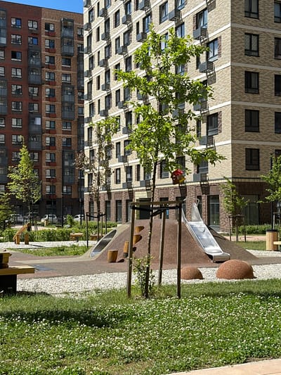 In the city of Znamya Oktyabrya, Russia, a modern residential complex is depicted. The foreground features a children's playground with a slide, wooden climbing structures, and decorative rounded red boulders. Lush green grass, dotted with small white flowers, covers the ground in the immediate foreground. In the midground, young trees with green foliage stand near the playground. Behind the playground, two multi-story residential buildings are visible. The building on the left is constructed with reddish-brown brick and features numerous windows and balconies. The building on the right, lighter in color with beige brick, also has many windows and balconies, and air conditioning units are visible on its exterior. The scene is bathed in bright sunlight, casting shadows on the buildings and playground structures. No people are present in the image.
