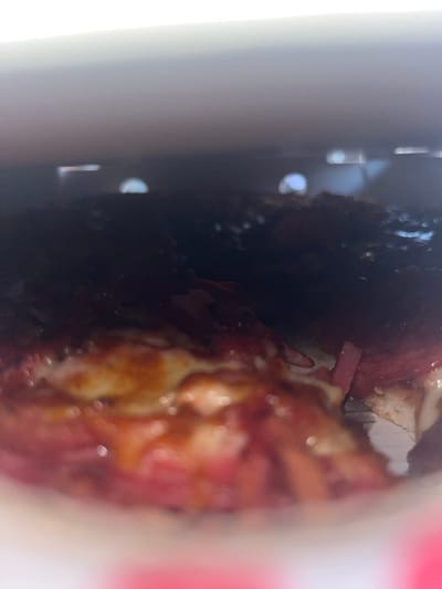 The image displays a close-up, low-angle view of the interior of a container, likely a pizza box, situated in Applecross, Australia. The central subject is a food item, consistent with a pizza, occupying the lower and mid-sections of the frame. The food features a visible crust, melted cheese with areas of browning, and numerous red-hued toppings, indicative of processed meats such as pepperoni or ham, interspersed with darker elements that appear to be sauce or other ingredients. The upper portion of the image is predominantly dark, suggesting the inside of a lid or the container's upper structure, against which several blurred, semi-circular and linear light reflections are discernible. The immediate foreground shows a white surface with a red pattern, characteristic of food packaging. No human figures or discernible actions are present in the static scene.
