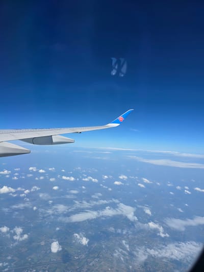 The image shows an aerial view of Linying County, China, from the window of an airplane. The airplane's wing, with a blue and red logo, is visible on the left side of the frame. Below, a patchwork of agricultural fields and some scattered buildings can be seen, partially obscured by a layer of white clouds. The sky is a clear, vibrant blue. The perspective suggests the plane is in flight at a considerable altitude.