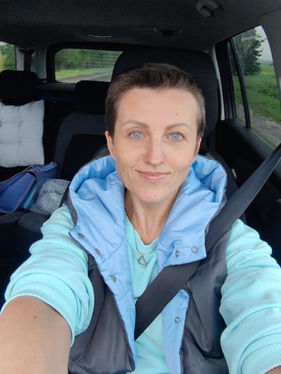 A woman with short brown hair and blue eyes is sitting in the driver's seat of a car. She is wearing a light blue hooded vest over a light blue sweatshirt and a silver necklace with a heart-shaped pendant. A seatbelt is fastened across her chest. Through the windshield and side windows, a rural landscape with trees and fields is visible, suggesting the car is on a road in a rural area, possibly near the Дзяржынскі сельскі Савет in Belarus, given the context. The interior of the car includes black car seats and a white pillow on the back seat.