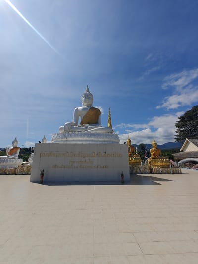 The image shows a large, silver-colored Buddha statue seated atop a platform inscribed with Thai text. The statue wears a golden robe. Several smaller, golden Buddha statues are arranged behind and to the right. The background features a bright blue sky with scattered clouds. A building with a traditional Thai roofline is visible on the right. The foreground is a wide, tiled courtyard. The inscription appears to dedicate the statue to a specific person and indicates the date of construction as December 27, 2557 in the Buddhist Era.