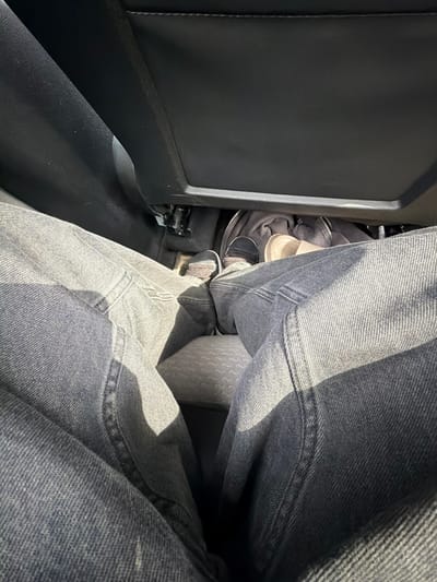 The image shows a first-person perspective of someone sitting in the back seat of a car. The subject is wearing dark blue jeans. In front of them, on the car floor, are a pair of dark sneakers with white soles, a cream-colored slipper, and a gray hooded jacket. The background consists of the dark leather seats of the car. The location is identified as Vallensbæk Landsby, Denmark.