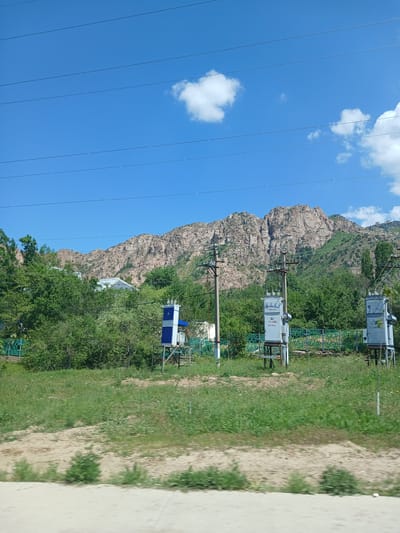 The image shows a landscape scene with a mountainous backdrop, lush green trees, and power infrastructure. The sky is clear blue with a few scattered white clouds and several horizontal power lines. In the foreground, there is a grassy field and a dirt path. Several electrical substations are visible along a green fence. Behind the trees, a building with a silvery roof can be seen. The mountains in the background are rocky and rugged, with sparse vegetation on their slopes. The scene appears to be a rural or semi-rural area. The prompt specifically mentions Chimgan, Uzbekistan, but this is not discernible from the image itself.