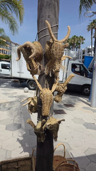 The image depicts a street scene in Almuñécar, Spain, where several woven straw animal heads, resembling bulls or donkeys, are hanging from a palm tree trunk. The foreground shows the base of the palm tree and a section of a paved sidewalk with a hexagonal pattern. In the background, a white delivery truck is parked, and a person is visible in the driver's seat. Beyond the truck, there are buildings with balconies and palm trees, suggesting a commercial or residential area. The sky is clear and blue, indicating a sunny day. Some woven baskets are also visible on the ground in the lower portion of the image.