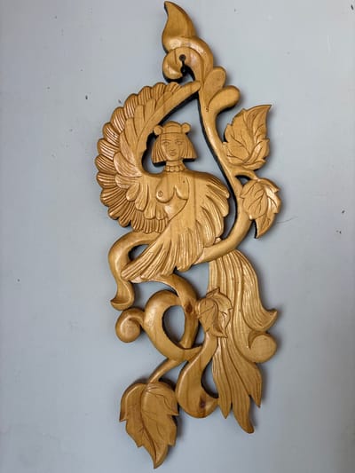 A wooden carving of a winged humanoid figure is mounted on a light blue wall. The figure has a human-like torso and head, with large, feathered wings extending from its back. Its lower body appears to be that of a bird or a stylized leg. The carving incorporates floral elements, with leaves and swirling vines integrated into the design. The wood is a light golden brown and appears to be varnished. The carving is vertically oriented and occupies the center of the frame. There are no people visible in the image. The location is identified as Аряж, Russia.