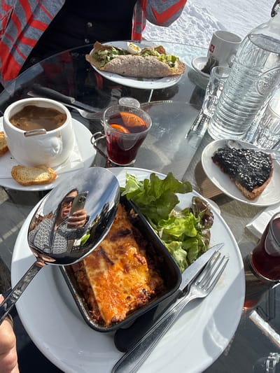 The image depicts an outdoor dining scene on a clear, sunny day in Saint-Martin-de-Belleville, France, with a snow-covered environment visible in the background. A round, dark, reflective glass table is occupied by various meal items and beverages.
**People:**
A person, identifiable by their reflection in a large, polished silver spoon held in the lower center foreground, is wearing sunglasses and a grey-patterned top, holding a smartphone. Another individual, wearing a grey and orange striped jacket, is partially visible seated on the left side of the table.
**Objects and Actions:**
The table is set with multiple dishes and drinks:
* In the upper left, a white plate holds a folded savory crepe containing green lettuce and topped with a slice of hard-boiled egg.
* To its right, a white ceramic cup shaped like an animal head contains a dark brown soup, accompanied by two pieces of toasted bread on a white napkin. A knife and fork rest behind this cup.
* A clear glass mug with a handle contains a red beverage garnished with orange slices.
* In the upper right background, a clear glass bottle filled with water, two empty clear glasses, and a white cup with a red logo on a saucer are present.
* In the lower right, a white saucer holds a slice of dark-colored tart with a spoon resting upon it. A glass with a red liquid is partially visible to the right.
* In the lower center, a rectangular black ceramic dish contains lasagna with a baked, golden-brown cheese topping, served on a white plate with a side of green lettuce salad. A fork and knife are positioned next to this plate.
The large spoon reflecting the person is held by a hand in the lower left foreground.
**Environment:**
The background features a bright, snow-covered ground or slope under direct sunlight. The scene is illuminated by strong natural light, causing reflections on the table surface and metallic objects.