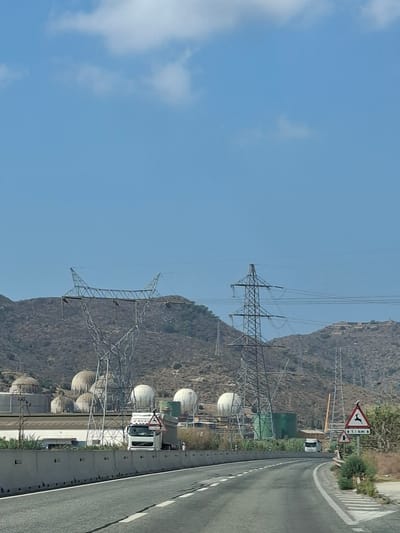 The image shows a section of a highway in Escombreras, Spain, with an industrial landscape visible to the left. The highway curves to the right, with a concrete barrier separating it from the industrial area. In the background, large spherical structures, possibly storage tanks, are prominent, along with several tall industrial towers and electrical pylons. A white truck with a trailer is driving on the highway. A road sign indicating a wildlife crossing is visible on the right side of the highway. The sky is blue with some scattered clouds. The terrain is hilly and arid.