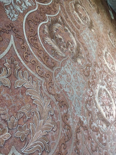 The image shows a close-up of a textured wallpaper with an ornate damask pattern in shades of brown and metallic silver. The pattern features scrolling floral motifs and elaborate curlicues. The lighting suggests the wallpaper is on a wall, possibly in a residential or commercial interior. There is no visible indication of the specific location within the city of Otaci, Moldova.