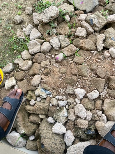 An overhead view shows a person's feet and legs wearing black sandals and a dark garment, placed on a pile of rough, gray and brown rocks and dirt. Small green weeds are scattered amongst the rocks. The scene is outdoors, possibly a construction or demolition site in Dumnga, Nigeria. The lighting suggests it is daytime.