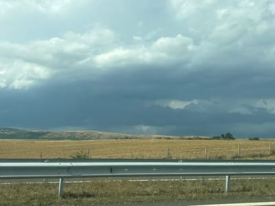 The image shows a cloudy sky over a dry, harvested field. In the foreground, a metal guardrail runs horizontally across the frame. The field is light brown and appears to be agricultural land, with some scattered green bushes and thin wooden posts. In the background, rolling hills are visible under the dramatic, dark clouds. The overall scene suggests a rural or semi-rural environment, possibly near Zheleznik, Bulgaria, given the context provided. The weather appears overcast, with the dark clouds suggesting the possibility of rain. No people or animals are visible in the image.