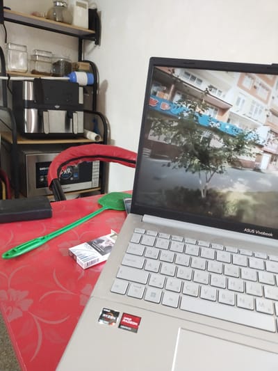 The image shows a close-up of a red table with a floral pattern. On the table are a pack of "MILANO" cigarettes, a green fly swatter, and a closed black object. A silver laptop with a visible keyboard and trackpad is positioned on the right side of the table. The laptop screen displays a video of an outdoor scene, possibly a street with buildings and trees. On the lower right corner of the laptop, there are stickers for "AMD RYZEN" and "AMD RADEON GRAPHICS". In the background, a shelf unit holds various items, including glass jars, a coffee machine, and a microwave. The scene appears to be indoors, with a plain white wall in the background. The location of Ikkiaul, Uzbekistan, is not directly indicated by any visual cues in the image.