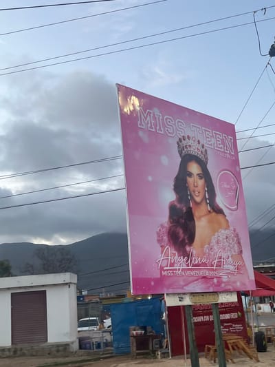 A large pink billboard advertising "MISS TEEN VENEZUELA" and featuring a crowned woman named Angelina Auaya is prominently displayed against a cloudy sky. The woman is wearing a sparkling crown and a voluminous pink dress. Below her name, it reads "MISS TEEN VENEZUELA 2025". The billboard is angled and appears to be located outdoors, with a mountainous landscape visible in the background. In the foreground, there are some low-rise structures, including a white building with a rolled-down metal door and a blue container-like structure. A red awning and some wooden tables and chairs are also visible, suggesting a commercial area or roadside establishment. Wires are strung across the sky above the billboard. The scene is set in Conuco Viejo, Venezuela.