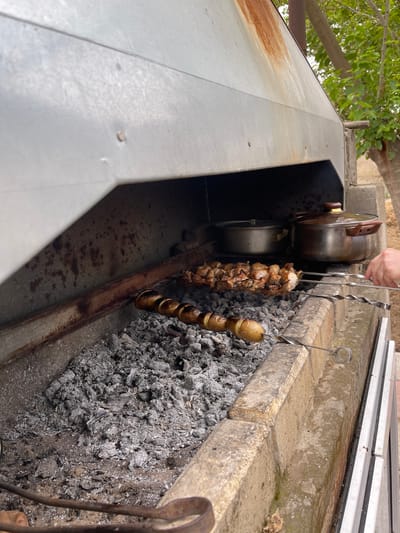 The image shows an outdoor barbecue area with a metal grill and brick base. A skewer of cooked meat is hanging over hot coals, and another skewer with potatoes is also present. There are metal pots or containers placed inside the grill. A person's hand is visible holding a pair of tongs. The scene suggests a meal being prepared outdoors, likely in a rural or suburban setting given the trees and foliage visible in the background. The lighting suggests it is daytime. There is no visible text in the image. The location context is Sabunchu Raion, Azerbaijan.