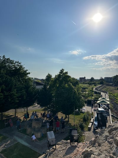 The image depicts a sunny day in Belene, Bulgaria, with a clear blue sky and scattered clouds. The sun is bright, casting long shadows, suggesting it is either morning or late afternoon. The scene appears to be an outdoor gathering or event taking place in a park-like setting, possibly near a historical or monumental area given the stone structures and the slight elevation from which the photo is taken.
Several people are visible, scattered throughout the scene. Some are standing in small groups, while others are sitting or lying down, seemingly relaxing or participating in an activity. One group is gathered around what appear to be large stone objects, possibly part of an exhibit or historical remnants. There are also trees and manicured greenery, suggesting a well-maintained public space. In the background, buildings and vehicles can be seen, indicating the presence of a town or city. A prominent white van and a lighter colored car are parked on a paved area. The overall atmosphere seems relaxed and leisurely. The image is oriented sideways, suggesting it was taken while the photographer was leaning or looking down from a higher vantage point.