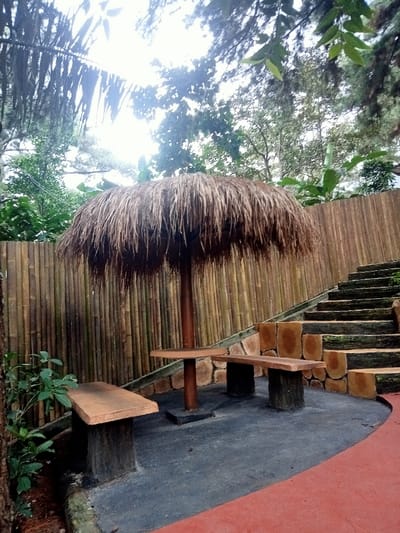 The image displays an unoccupied outdoor seating area within a verdant setting in Kuwukan, Indonesia. A central, circular thatched umbrella with a dark brown, fibrous roof is supported by a single dark pole. Beneath it, a light brown circular table with a central pedestal base is flanked by two rectangular benches, each with a light brown top and dark, block-like supports. These objects rest on a dark grey paved surface that curves towards a reddish-orange pathway in the lower right foreground. Behind the seating area, a tall, light brown wall constructed from numerous vertical bamboo poles spans the background. To the right, a set of ascending stairs, formed from stacked, horizontally oriented log-like concrete elements, rises alongside the bamboo wall. Lush green foliage, including tree canopies and smaller plants, is visible above the bamboo wall and in the upper left, with an overhanging large leaf partially obscuring the sky. No individuals or living creatures are present in the scene.