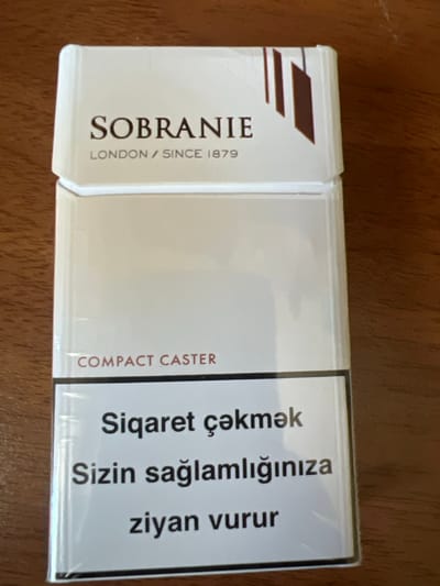 The image shows a cigarette pack of the brand "SOBRANIE" with "LONDON / SINCE 1879" printed below. The text "COMPACT CASTER" is visible on the lower part of the pack, followed by a warning in a foreign language, which translates to "Smoking harms your health". The cigarette pack is placed on a wooden surface. The lighting suggests it is an indoor environment, possibly a home or office setting. No specific location in the city of Sabunchu, Azerbaijan is indicated by any visible elements in the image.