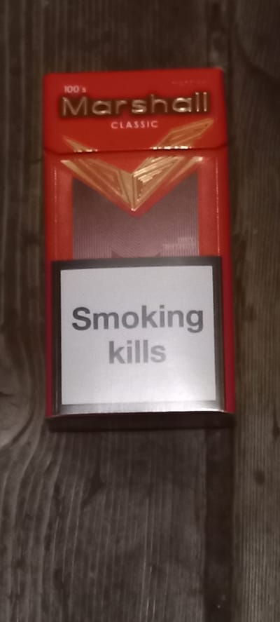 A red cigarette pack, branded "Marshall Classic 100's", lies on a dark, wooden surface. The pack has a golden geometric design on its upper half. The bottom half of the front of the pack features a white label with the warning text "Smoking kills" in grey lettering. The image is a close-up, vertical shot of the cigarette pack. There are no people or other objects visible in the scene. The location in Markevycheve, Ukraine, is not discernible from the image itself.