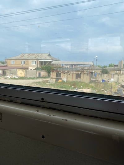 This image, taken from a window, looks out onto a cloudy sky and a scene of modest buildings and overgrown vegetation in Novy Khushet, Russia. The foreground shows the interior of a vehicle or building, with a beige-colored surface and what appears to be a textured wall below. The window itself is dirty and scratched, with reflections of overhead wires visible.
The view outside reveals several low-rise buildings, some with visible damage or incomplete construction. One prominent building in the middle ground has a light-colored, slightly sloping roof and appears to be made of brick or concrete. Adjacent structures are also visible, some with corrugated metal roofing. The ground is covered in sparse, tall grass and scattered debris, suggesting an unkempt or developing area. A lamppost is visible in the distance. The overall impression is one of a somewhat rural or semi-developed urban periphery.