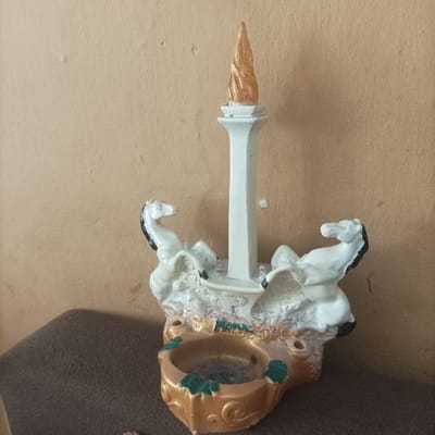 The image displays a decorative souvenir or functional item, possibly an ashtray or incense holder, positioned indoors in Pasir Muncang, Indonesia. The central object is a miniature replica of the Monumen Nasional (Monas) from Jakarta. This replica features a tall, slender white column topped with a golden, flame-like spire. Flanking the column are two rearing white horses, each with black manes and tails. The base of the object is multi-tiered and textured, primarily in shades of brown and gold, with distinct green leaf-like embellishments. A circular depression at the front of the base contains dark, granular residue, consistent with ash. The object is resting on a dark brown, possibly fabric, surface, against a plain, light brown or beige wall. No individuals are present in the scene.