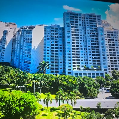 The image depicts a daytime aerial view of a modern urban landscape dominated by a series of tall, light-colored apartment buildings. The buildings are numerous and appear to be connected or in close proximity, forming a significant complex. They feature repetitive patterns of windows and balconies. In the foreground and surrounding the buildings, there is lush greenery, including numerous trees and palm trees, indicating a tropical or subtropical climate. Some of the buildings have terraces or rooftop amenities, such as lounge chairs and umbrellas, suggesting residential or recreational spaces. A street is visible in the lower right portion of the image with a few cars parked. The sky is clear and blue with a few scattered white clouds. This scene is not located in Lippstadt, Germany, which is in a temperate climate and would not have this type of dense, tropical vegetation.