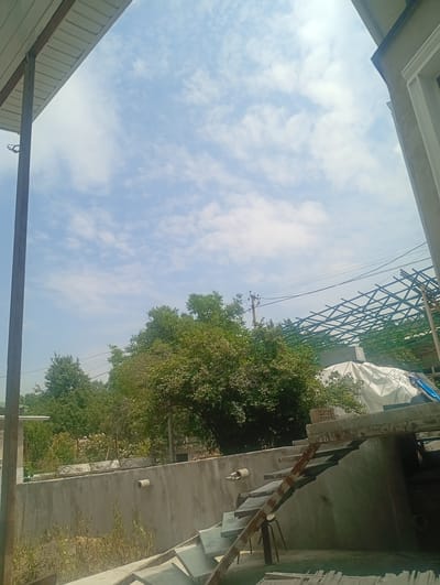 The image depicts an outdoor scene under a cloudy sky, with a view of Arashan, Kyrgyzstan. In the foreground, a concrete wall runs horizontally, with dried vegetation growing at its base. To the right of the wall, a set of grey concrete stairs with rusted metal railings ascends. Above the wall, a dense canopy of green trees is visible, with a few blooming pink flowers. A metal framework, possibly for a roof or pergola, is under construction in the background, along with a large white tarp. Power lines are strung across the sky, supported by utility poles. On the left side of the image, a portion of a building with a white roof and a dark metal support beam is present. The overall impression is of a rural or semi-constructed area in a natural setting.