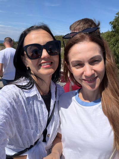 Two women are taking a selfie outdoors on a sunny day. The woman on the left has dark hair, is wearing large black sunglasses, and a white button-up shirt. The woman on the right has long, reddish-brown hair and is wearing a white t-shirt with a blue collar. Behind them, a man with short hair is visible, and further in the background, there are trees and a clear blue sky. The location appears to be a coastal area, possibly near Morskoye, Russia, given the vegetation and the presence of the sea in the distance. The overall scene suggests a casual outdoor activity.