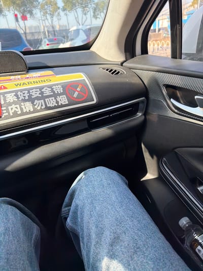 The scene depicts the interior of a vehicle, viewed from the front passenger seat towards the dashboard and exterior. The location is described as Jianye District, China. A person's legs, clad in light blue denim jeans, are visible in the lower foreground, indicating a seated individual. The black dashboard features horizontal air vents and a prominent yellow sticker with "WARNING" in English and Chinese characters reading "系好安全带" (Fasten seatbelt) and "内请勿吸烟" (No smoking inside), accompanied by a red circular no-smoking symbol. A small, dark-colored bottle with a black cap is situated in a compartment on the black passenger door panel, which also has a textured, patterned insert and a silver door handle. Through the front windshield, a linear crack is visible, and the exterior environment shows several other vehicles, bare trees, and a clear blue sky. A light-colored building with arched openings is visible in the distant background through the side window. The overall lighting suggests a sunny daytime setting.