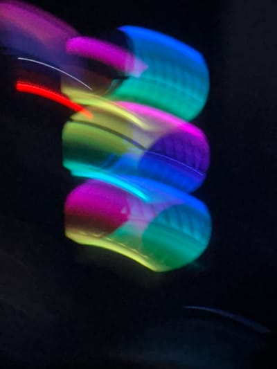 The image displays a long exposure photograph of neon lights in a dark environment, possibly a night scene. The lights are blurred and streaked due to motion, creating a vibrant and abstract display of colors including pink, yellow, green, and blue. The arrangement of the lights suggests they might be part of a sign or a decorative installation. The background is predominantly black, indicating low light conditions. There are no visible people, specific landmarks, or identifiable objects that would allow for precise location identification within Stallings, United States of America. The scene is purely abstract and focused on the visual effect of the moving lights.