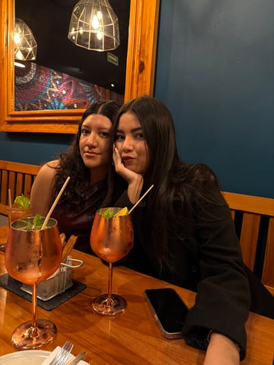 Two young women pose at a wooden table. The woman on top wears a black jacket, resting her head on her hand. Her dark hair flows down. Below, the other woman, with curly hair, smiles. Two copper-colored stemware glasses with straws and garnishes sit on the table along with a smartphone, silverware, and a dark rectangular object. The table and wooden chairs are set against a blue wall. A wood-framed mirror reflects light fixtures and an abstract art piece.