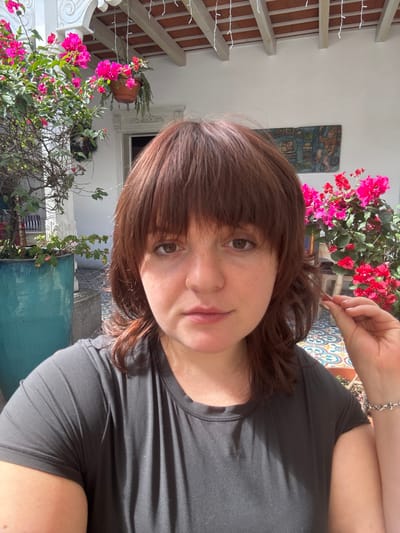 A woman with brown, fringed hair is centered, wearing a black T-shirt. She's indoors, with an outdoor courtyard visible behind her. Red and pink bougainvillea flowers are prominently displayed, along with a tile mural on the wall. A pergola with string lights extends along the building facade. A window is partially visible in the background. The woman is looking directly at the camera, and part of her arm and bracelet is visible at the top-right corner.