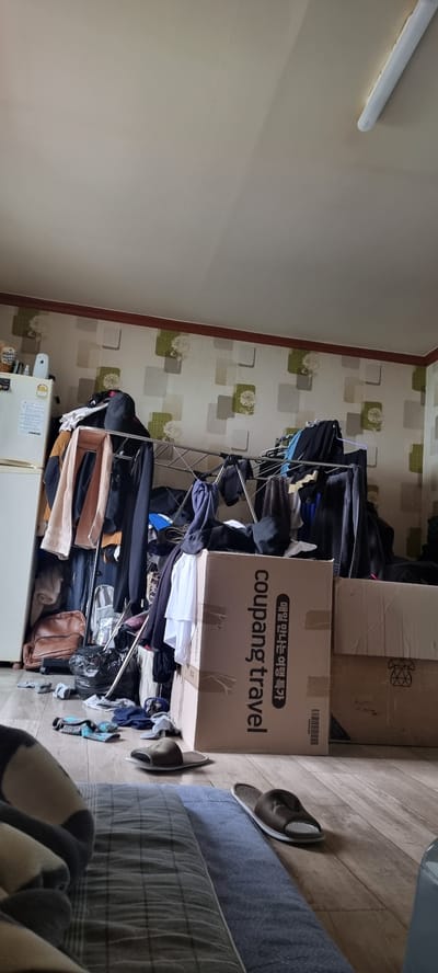 The image depicts a cluttered room, likely a bedroom or storage area, with various items strewn about. A drying rack, overloaded with clothing, dominates the center of the frame. Next to it, a large cardboard box with "coupang travel" printed on it is visible, along with other boxes and bags. A white refrigerator stands in the lower left corner. On the floor, a pair of sandals rests near a grey blanket, and a pair of socks is also visible. The room has patterned wallpaper and wooden flooring. The lighting suggests it is daytime. The overall impression is one of disarray and a lack of organization. There is no visible indication of people or any specific activity taking place beyond the presence of stored items. The text "coupang travel" is clearly legible on the box. The location is identified as Jinju-si, South Korea.