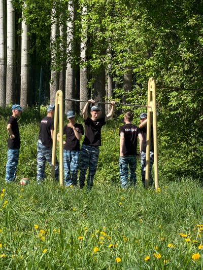 The image shows four individuals wearing black shirts with the word "ПОЛИЦИЯ" (POLICE) printed on them and camouflage pants. They are standing in a grassy area with dandelions. The individuals appear to be performing exercises on a metal structure, possibly a fitness station in a park or training ground. The background includes trees and foliage, and a birch fence. A volleyball is on the ground. It is daytime, with bright sunlight illuminating the scene.
While the description provides a visual account of the scene, it's impossible to pinpoint the exact location within the city of Oreshniki based solely on the image.