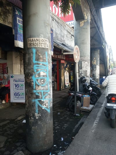 A daytime street scene shows two gray, cylindrical support pillars, with a shop on the left. Graffiti is visible on the left pillar along with a sticker advertising "PASANG CCTV 081806707074". A person sits on a red stool next to a promotional sign for "FlexMax XL" voucher packs. Two motorcycles are parked next to the right pillar, with another motorcycle driving on the street. A woman wearing a white jacket is seen riding the last motorcycle. A traffic sign with a crossed-out figure is mounted on the second pillar. The shopfront displays mannequins with clothing.