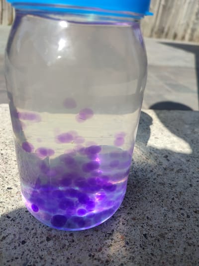 A clear plastic jar filled with water and purple water beads sits on a concrete surface outdoors. The jar has a blue lid and appears to be partially filled, with the water beads concentrated at the bottom and scattered throughout the lower portion of the liquid. Sunlight casts shadows on the concrete. The background shows a blurred wooden fence. The location is identified as Velykyi Zhytyn, Ukraine.