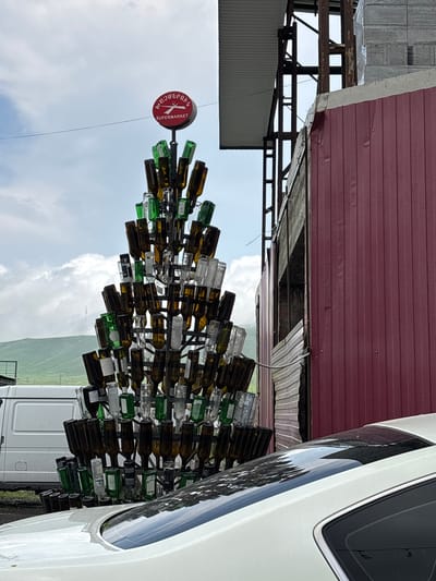 In Tsovagyugh, Armenia, a roadside supermarket is marked by a towering, unconventional Christmas tree constructed entirely from glass bottles. The tree, positioned in front of a building with a deep red corrugated metal facade, is a multi-tiered structure of brown and green bottles, some clear, arranged to resemble branches and foliage. A circular red sign with white text, reading "SUPERMARKET" and some text in Armenian script above it, crowns the bottle tree. To the left of the bottle tree, a white van is parked. In the foreground, the rear section of a white car is visible, its roof reflecting the cloudy sky and surrounding greenery. The background features rolling green hills under a partly cloudy sky, with a faint, distant structure resembling a tower on one of the hills. The construction site to the right of the building appears to be in progress, with exposed metal beams and stacked building materials.