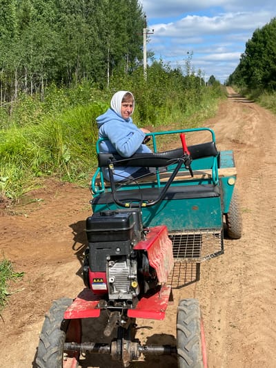 A woman wearing a light blue hooded sweatshirt and a white headscarf sits on a green trailer attached to a red and black tiller. The tiller has a prominent engine and two large wheels. The trailer has two seats, the one the woman is occupying is black and padded, and there is a red lever on the side. The trailer also has a green metal frame. The tiller and trailer are on a dirt road. Tall green grass and trees line the sides of the road. The sky is blue with white clouds. The location is identified as Dresvyanovo, Russia.