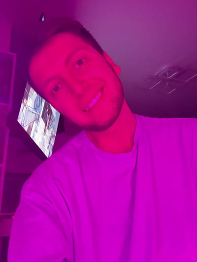 A young adult male is captured in a close-up selfie with a tilted head, bathed in bright pink lighting. He has short brown hair, a light beard, and is wearing a plain t-shirt. His eyes are open, and he is smiling, revealing white teeth. The background is indistinct due to the lighting, but a television screen is visible to his left, displaying an image of buildings. A ceiling fan and part of a light fixture are also discernible in the upper right portion of the frame. There is no information in the image to suggest the location is Tokaryovo, Russia.
