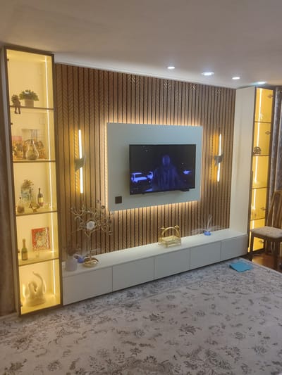The image depicts a living room in Chuy, Kyrgyzstan, featuring a modern entertainment unit against a backdrop of vertical wooden slats. A large flat-screen television is mounted on the wall. To the left, a glass-fronted display cabinet with internal lighting showcases various decorative items, including figurines and framed photographs. To the right, a similar display cabinet is visible, along with a wooden chair and curtains. The floor is covered with a patterned rug. The lighting in the room appears to be a mix of recessed ceiling lights and the warm glow from the display cabinets and wall-mounted sconces. The overall ambiance is one of a well-lit and contemporary living space.