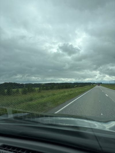 The image depicts a view from the front of a vehicle traveling on a paved road. The road stretches into the distance, with a white line marking the right edge of the lane. To the left of the road is a grassy field interspersed with trees and a dense line of dark green trees forming a forest in the background. The sky above is overcast with heavy, grey clouds. Two trucks are visible on the road ahead, traveling in the same direction as the camera. The foreground shows the dashboard and windshield of the vehicle, with reflections on the glass. The overall scene suggests a rural or semi-rural setting, likely during daytime under a cloudy sky. There is no specific indication of the city of Kostovaty, Russia, in the image.