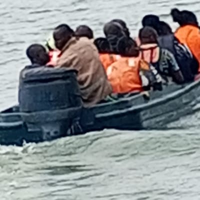 A dark-colored open motorboat is depicted traversing choppy water near the city of Koko, Nigeria. The vessel contains at least ten individuals. An operator, clad in dark clothing, is positioned at the stern, managing a dark-colored outboard motor. Among the passengers, a minimum of five individuals are wearing bright orange life jackets. Other passengers are dressed in civilian attire, including one person in a light brown jacket and another in a patterned shirt. The boat is actively in motion, generating a visible wake on the water surface.