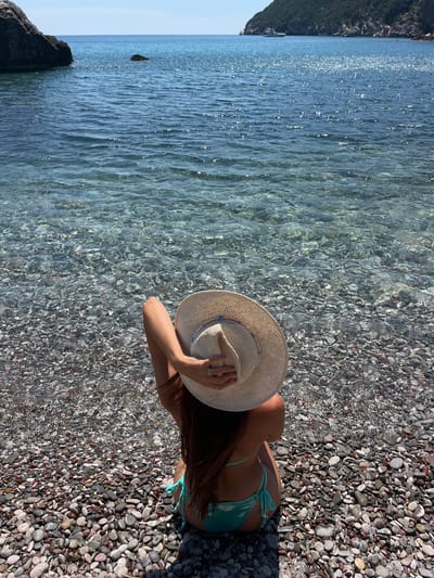 The image shows a person sitting on a pebble beach next to clear, shallow water. The person is wearing a light-colored straw hat and a turquoise bikini. Their back is to the camera, and they are holding the hat with one hand. The water is calm and reflects the sunlight, creating shimmering patterns on the surface. The beach is covered in smooth, gray and brown pebbles. In the background, the sea extends to the horizon, and a rocky, tree-covered hill is visible on the left side of the frame. The lighting suggests it is daytime, with bright sunlight. The scene evokes a sense of relaxation and enjoying a day at the beach. There is no visible text in the image.
