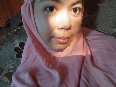 The image is a selfie taken indoors. The subject is a young woman wearing a pink hijab and a pink top. She is looking directly at the camera with a slight smile. Her face is illuminated by a bright light source, casting a shadow across her left cheek. The background shows a textured couch and a wooden surface, possibly a table or floor. The scene does not contain any elements that would indicate its location in Labissa, Indonesia.