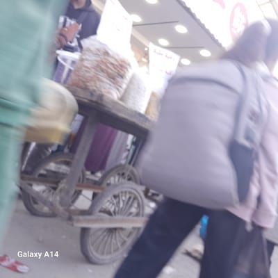 The image appears to depict a street scene in Al Mawasi, with a mobile cart laden with bags of goods, possibly food items, positioned in the foreground. The cart has a weathered, wooden frame and visible spoked wheels. Behind the cart, two individuals are walking. A person with a large backpack is partially obstructing the view of another person wearing a pink garment. The background features what seems to be a storefront with visible signage, including one with possible Arabic text and another with a circular logo with pink coloration. A partially visible person with a bucket can be seen behind the cart. The image is blurred, suggesting motion. The bottom of the image shows "Galaxy A14," indicating the camera used.