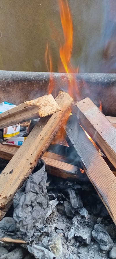 This image captures a close-up view of a fire burning through wooden planks and what appears to be crumpled paper or fabric. The fire is intensely bright, with vibrant orange and yellow flames licking upwards and smoke curling around the burning materials. Several pieces of wood, some still relatively intact and others charred and broken, are arranged in a way that suggests they are fueling the fire. The background on the left is a textured, muted surface, possibly a tarp or wall, with the flames reflecting on it. The right side of the image is dominated by piles of dark ash and burnt material, indicating the fire has been burning for some time.
The scene is chaotic and dynamic due to the active flames and smoke. There are no people visible. The focus is entirely on the destructive process of the fire. The overall impression is one of heat and consumption. No specific time of day or weather conditions can be inferred from the image.
Visible text is minimal and partially obscured. On a piece of what looks like packaging tucked amongst the burning material, the word "KAR" is faintly visible, followed by a series of numbers that appear to be "0200 0 10 223". Due to the angle and burning, the text is not fully legible. The location context of Iskrets, Bulgaria, does not provide any direct visual cues within the image itself.