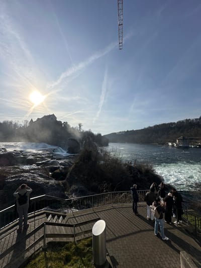 The image depicts the Rheinfall waterfall in Neuhausen am Rheinfall, Switzerland, under clear, sunny conditions. The bright sun is positioned in the upper left quadrant of the light blue sky, which is marked by multiple intersecting contrails forming an "X" pattern. The metal jib of a large construction crane is visible extending from the upper right corner of the frame.
In the midground, the powerful Rheinfall cascades over rocky terrain on the left, generating a significant amount of mist that partially obscures Schloss Laufen, a prominent castle structure situated on the hill overlooking the falls on the left bank. Downstream, the Rhine River flows from left to right, exhibiting dark, choppy water. A multi-story building with white facades and dark roofs, likely a hotel or restaurant, is visible on the far bank of the river in the right midground, surrounded by dense, leafless trees.
In the foreground, an observation platform, paved with grey interlocking pavers and enclosed by a dark metal railing, occupies the lower portion of the image. A set of wooden steps with metal railings leads down to this platform from the left. On the left side of the platform, one individual, viewed from behind, wears a light-colored jacket and a backpack, standing with hands raised towards their head, observing the waterfall. On the right side of the platform, a group of approximately 6-8 people is gathered near the railing, also looking out at the falls and river. They are dressed in casual winter attire. A cylindrical, silver-colored metal post stands on the platform to the right of the staircase. The surrounding ground near the platform shows patches of dry grass and dark, bare vegetation.