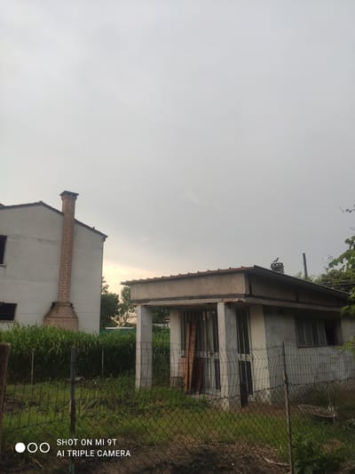 The image depicts an outdoor scene in Bagnoli di Sopra, Italy. The primary subjects are two houses and a fence. One house appears to be under construction or renovation, with exposed concrete and rebar, and windows that are either boarded up or have bars. It has a covered porch area supported by columns. The other house is partially visible and features a prominent brick chimney. Lush green vegetation, possibly crops or tall grass, is visible behind the fence. The sky is overcast, suggesting a cloudy day or possibly early morning or late afternoon. The overall atmosphere is one of rural or semi-rural residential development.
Visible text on the lower right of the image reads: "SHOT ON MI 9T AI TRIPLE CAMERA". This indicates the image was captured using a Xiaomi Mi 9T smartphone. There are no people visible in the frame, and no specific activity or event is taking place; it appears to be a still shot of the environment. The lighting suggests it is not midday, but the exact time is difficult to determine due to the overcast conditions.