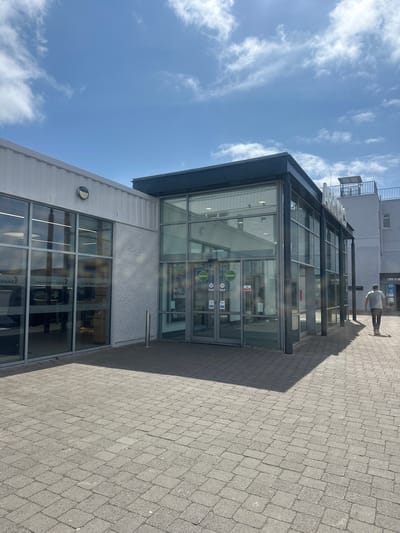 The image shows the exterior of Ireland West Airport Knock, located near Charlestown, Ireland. The building features a modern design with large glass windows and a grey facade. The foreground is paved with grey interlocking bricks. A person is walking away from the entrance on the right side of the frame. Above the entrance, large white letters spell out "ARRIVALS." The sky is blue with scattered white clouds. The overall scene depicts a bright, sunny day outside the airport.